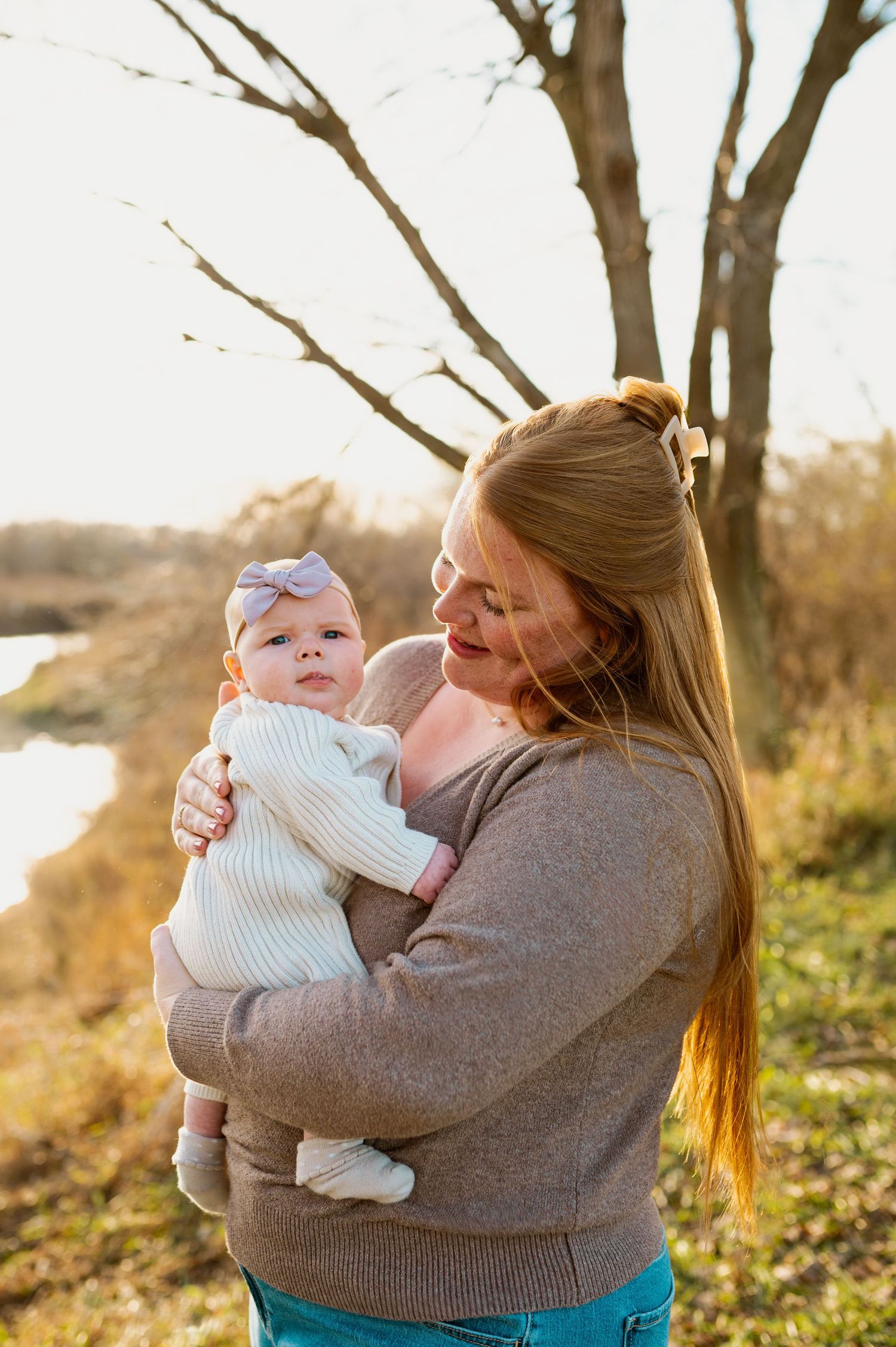 A woman in a gray sweater holds and plays with a baby outdoors during golden hour in autumn.