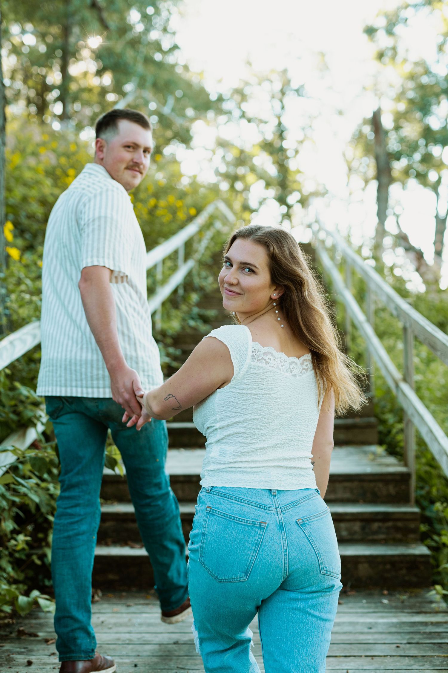 A couple poses on outdoor wooden stairs surrounded by lush greenery on a sunny day.