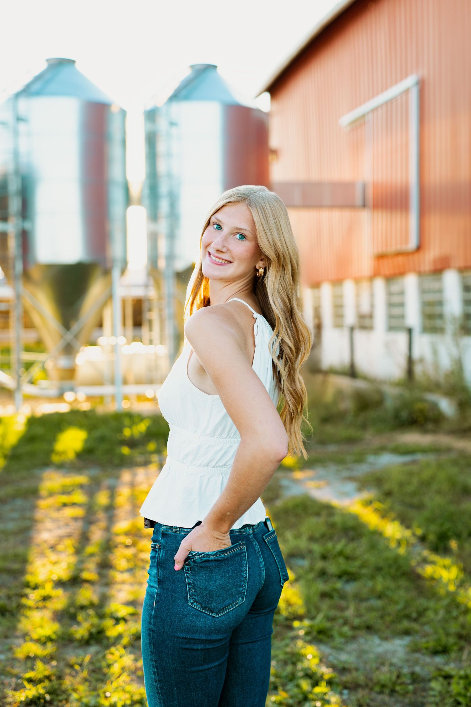 A woman in white top and jeans poses against a rustic barn backdrop in warm sunset lighting.