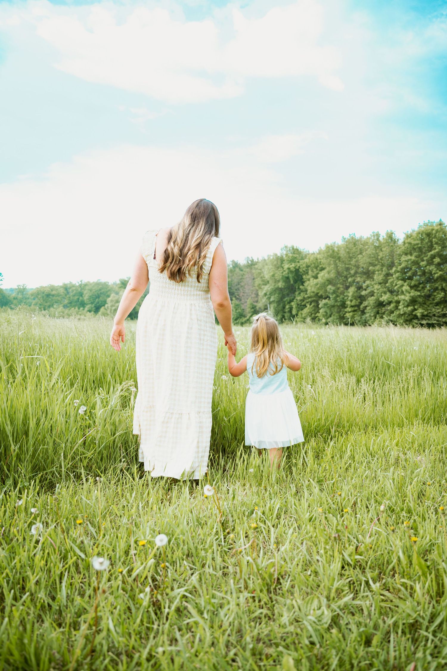 Two figures in white dresses walking through tall summer grass field on sunny day.