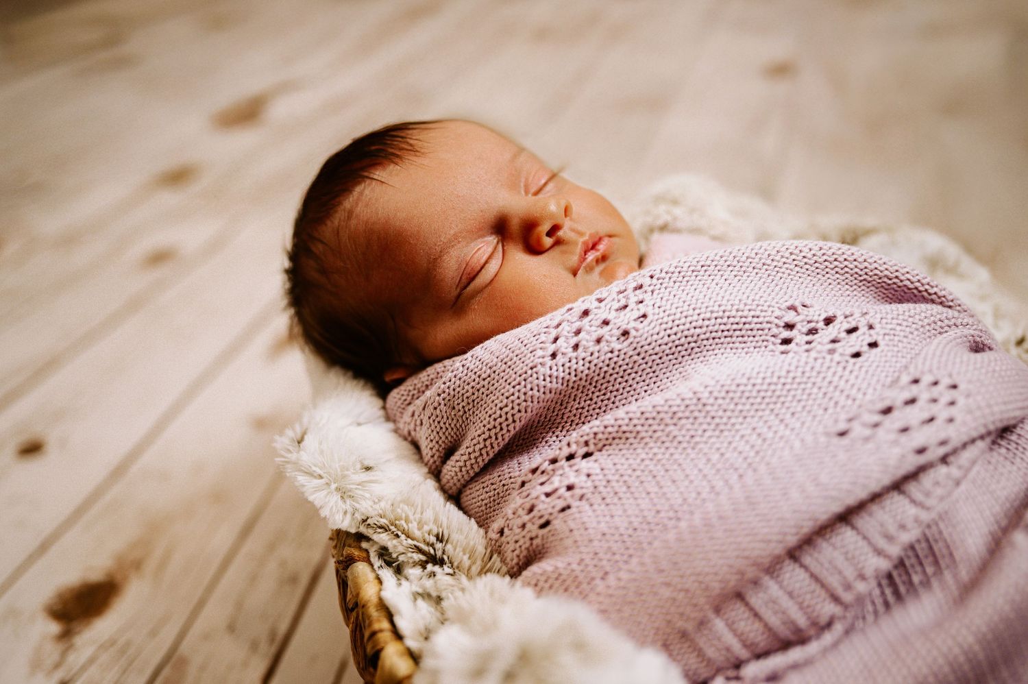 Sleeping newborn baby wearing pink knit sweater resting on wooden floor.