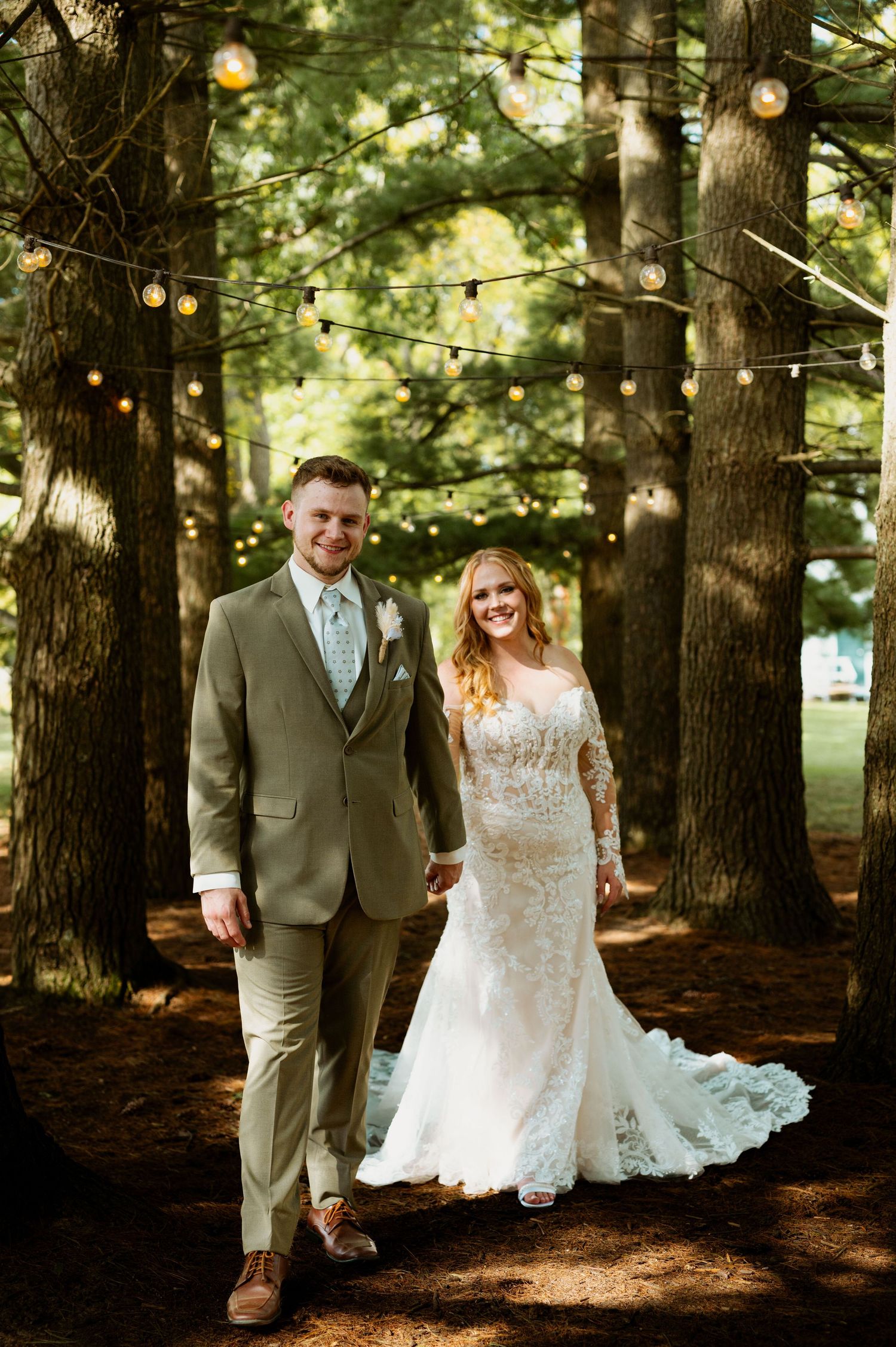 Newlyweds walk hand in hand through a forest path decorated with string lights at dusk.