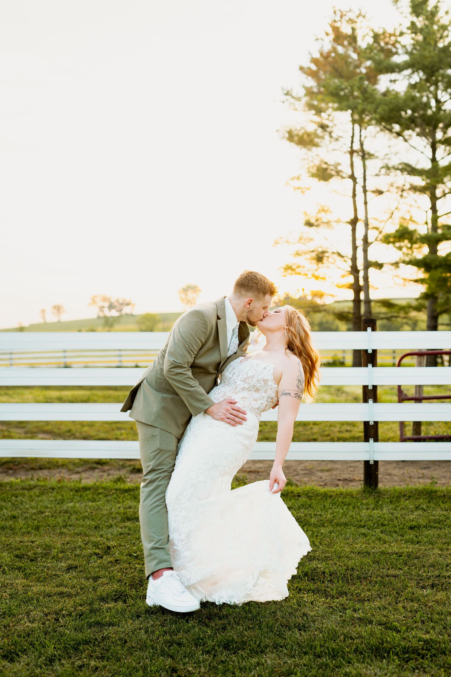 Romantic wedding couple sharing a tender moment on grass with white fence and sunset backdrop.
