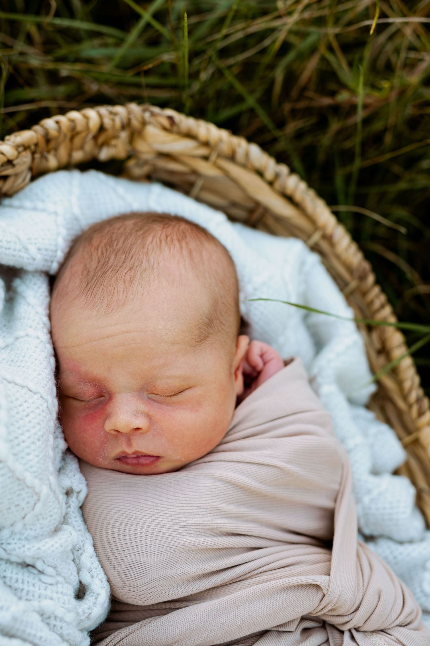 Sleeping newborn baby wrapped in beige swaddle blanket and white towel lying in wicker basket.