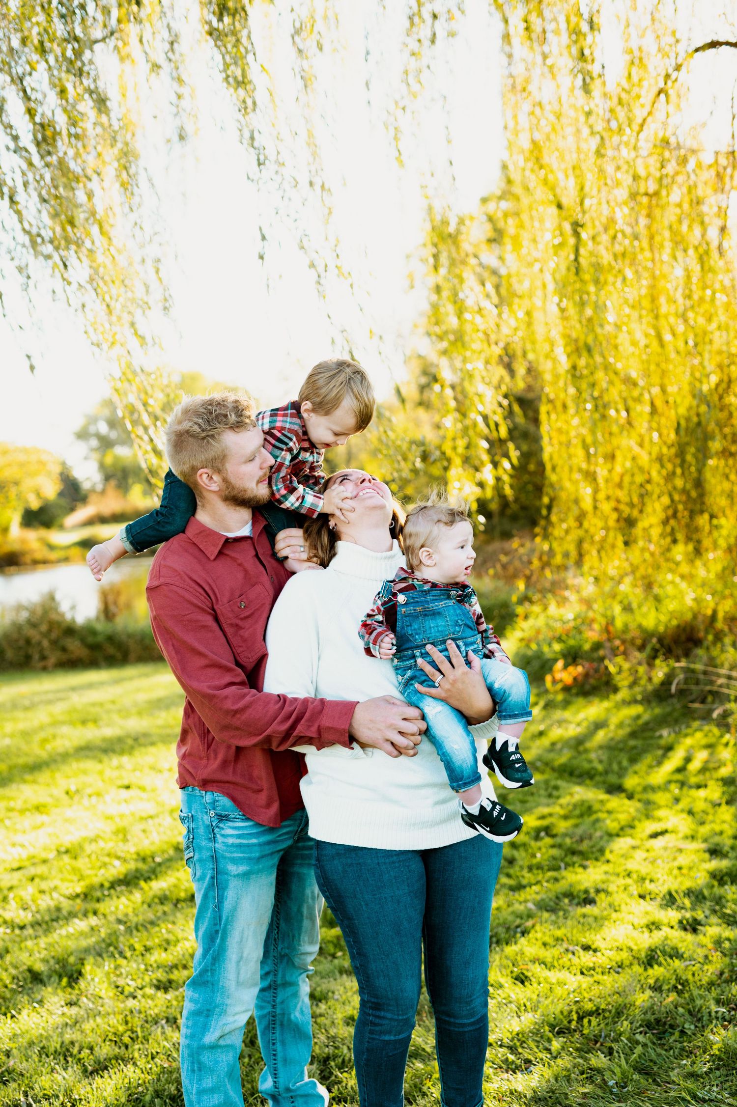 A family shares playful moments together in a park setting with weeping willow trees in the background.