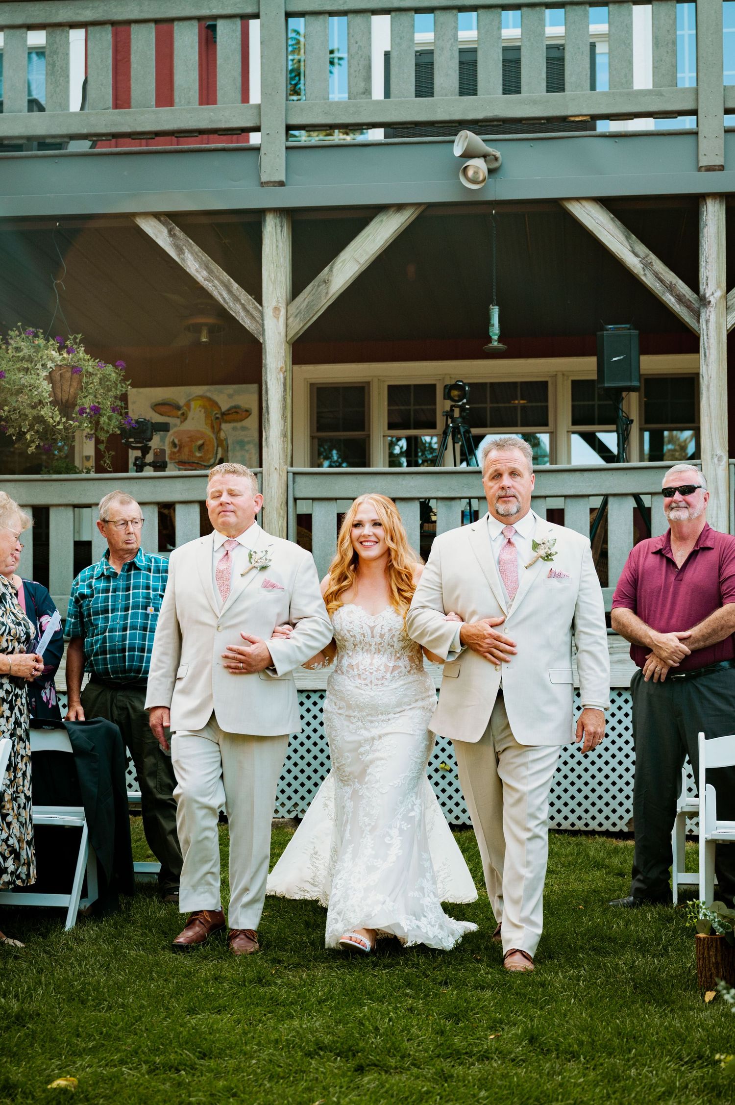 A bride in a white lace dress walks down a grassy aisle escorted by two men in light colored suits.
