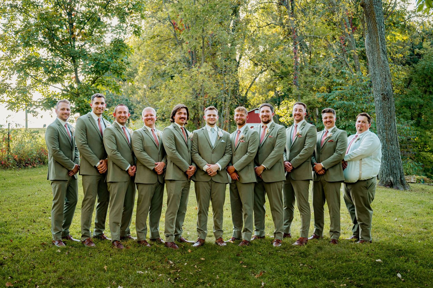 Wedding party in sage green and neutral attire poses in a line against a natural outdoor backdrop.