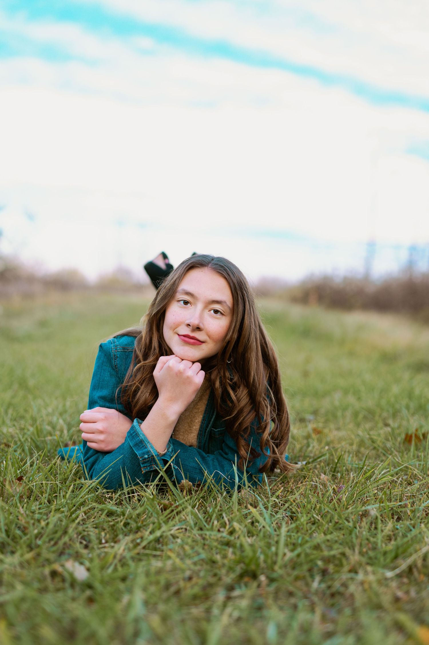 A person in a teal sweater lies in grass during a sunny outdoor photo session.