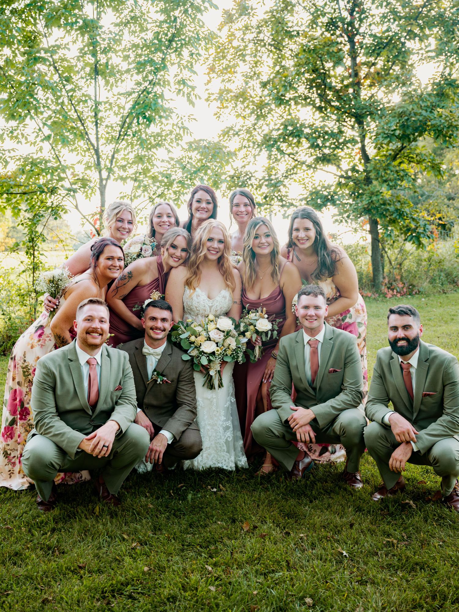 Large wedding party group photo with bridesmaids and groomsmen sitting on grass.