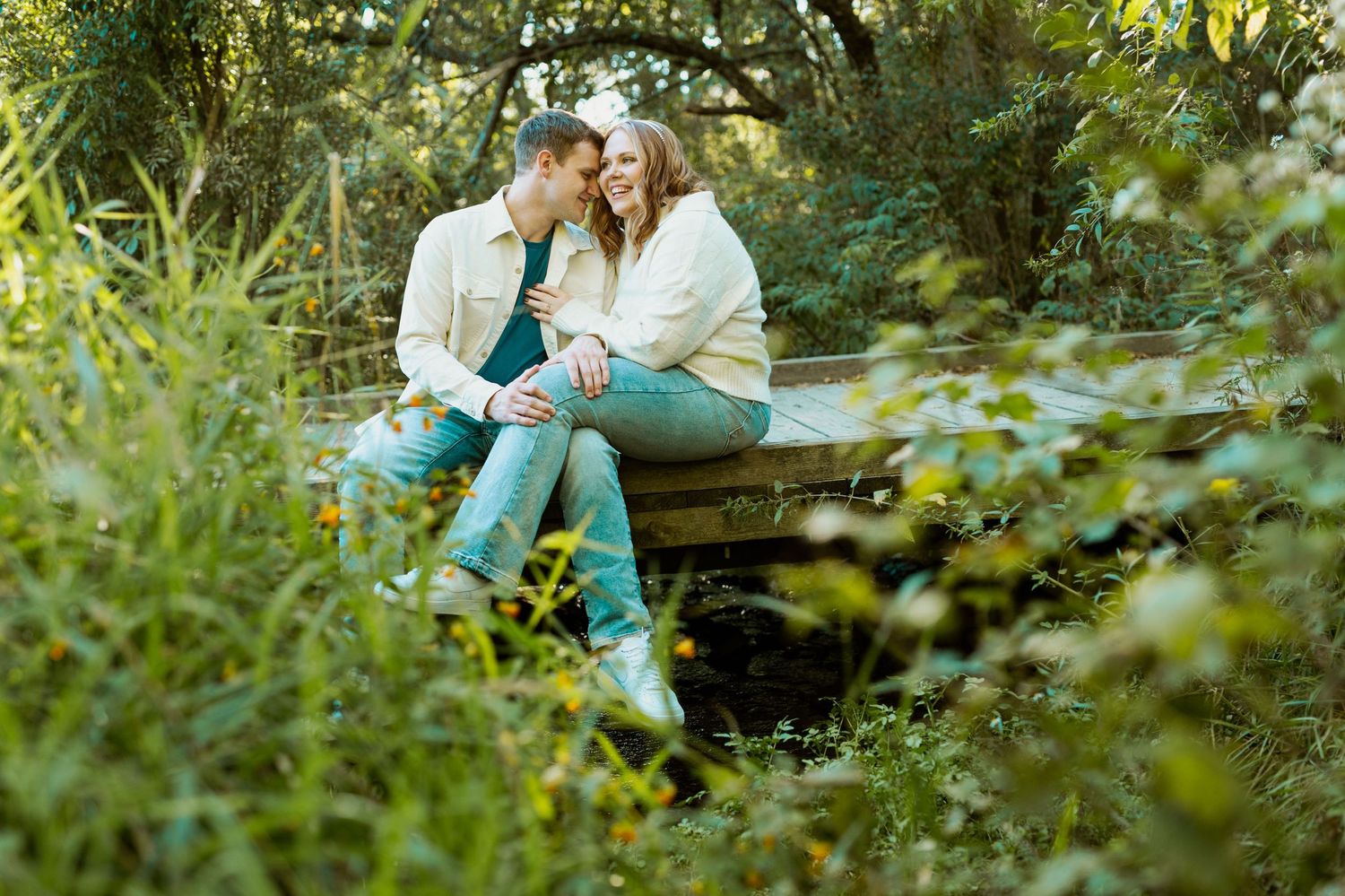A couple sits together on a wooden bridge surrounded by lush green foliage.