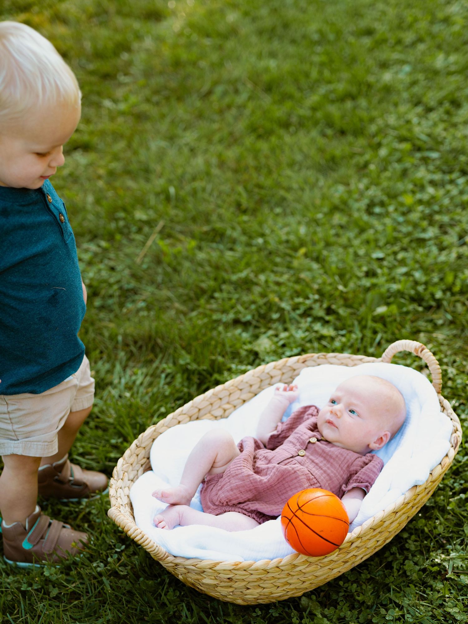 A baby lying in a woven basket on grass with a small orange basketball nearby.