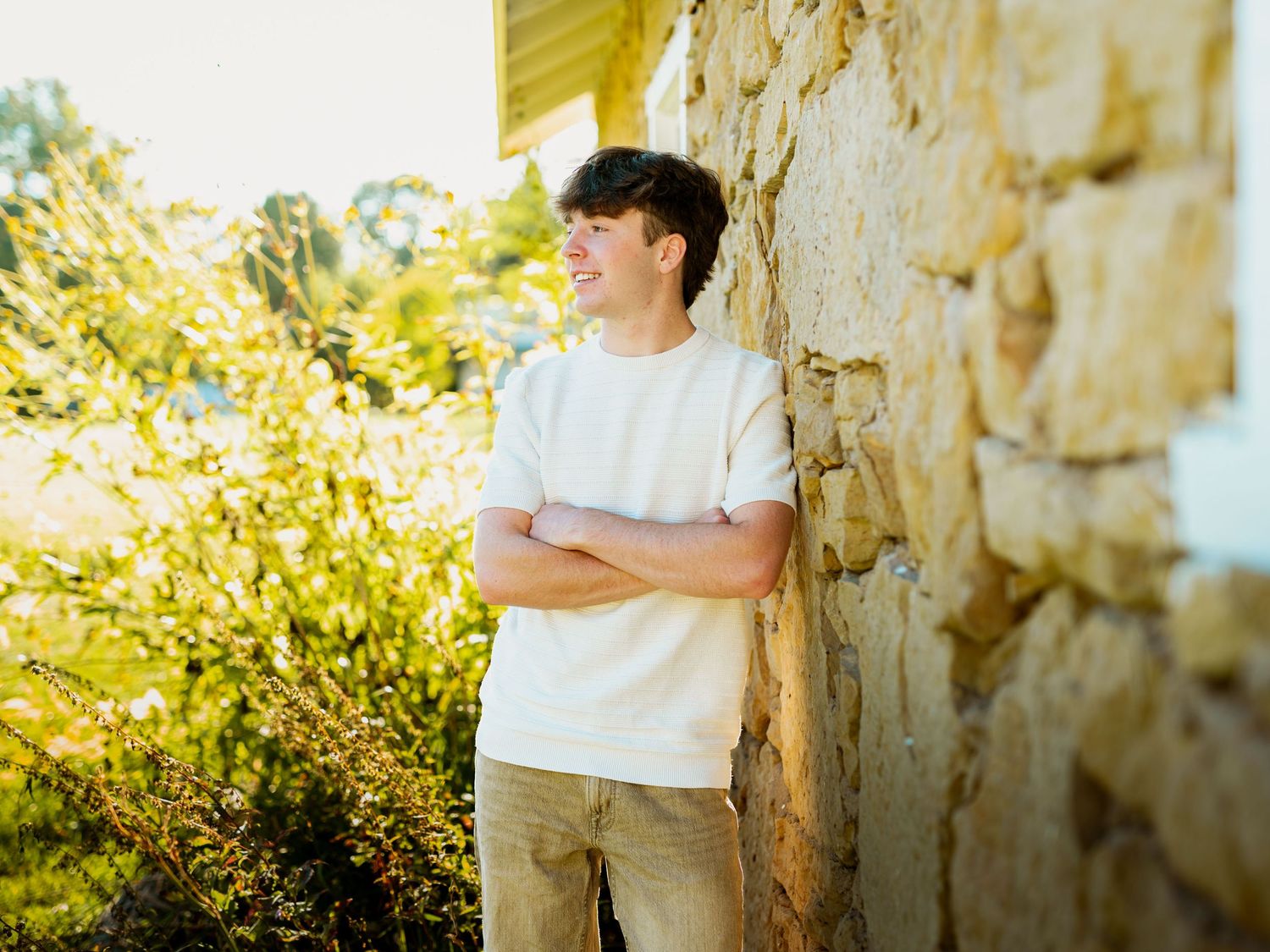 A couple in light colored clothing embraces in front of a rustic stone wall on a sunny day.