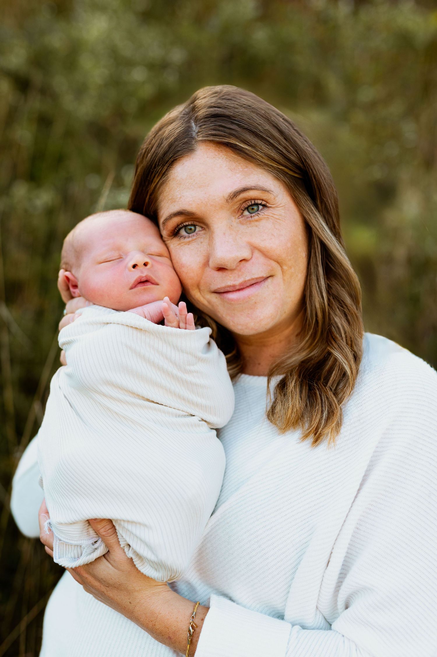 A woman in a white sweater shares sweet moments with a newborn baby in an outdoor setting.