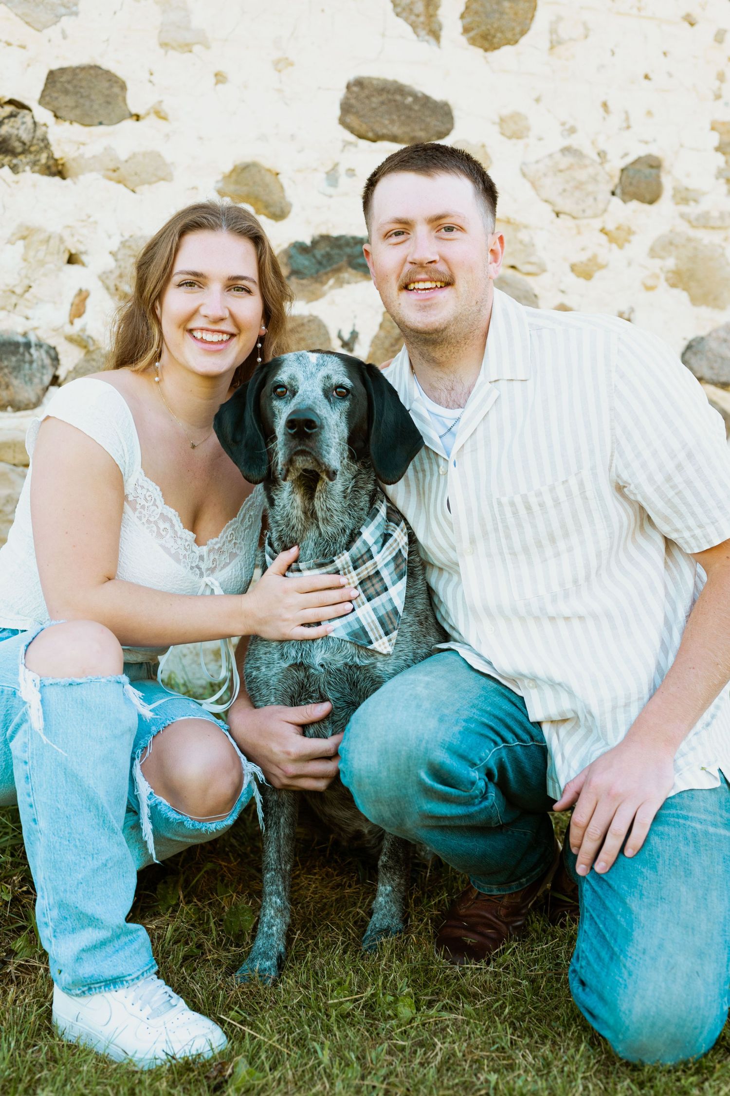 A happy couple poses with their black dog against a rustic stone wall background.
