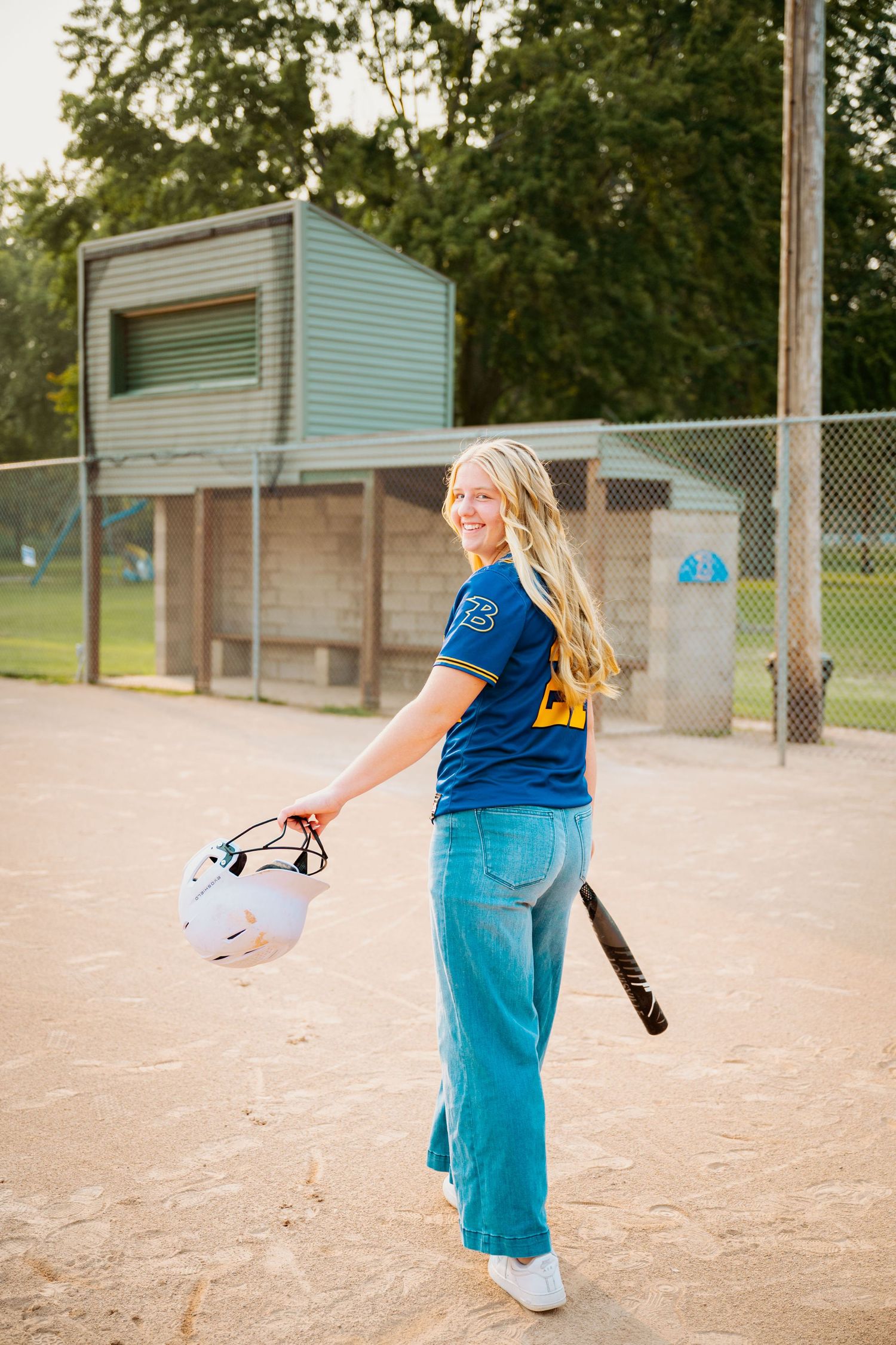 Softball player in blue jersey and teal pants holding a helmet while standing on a dirt field near dugout.