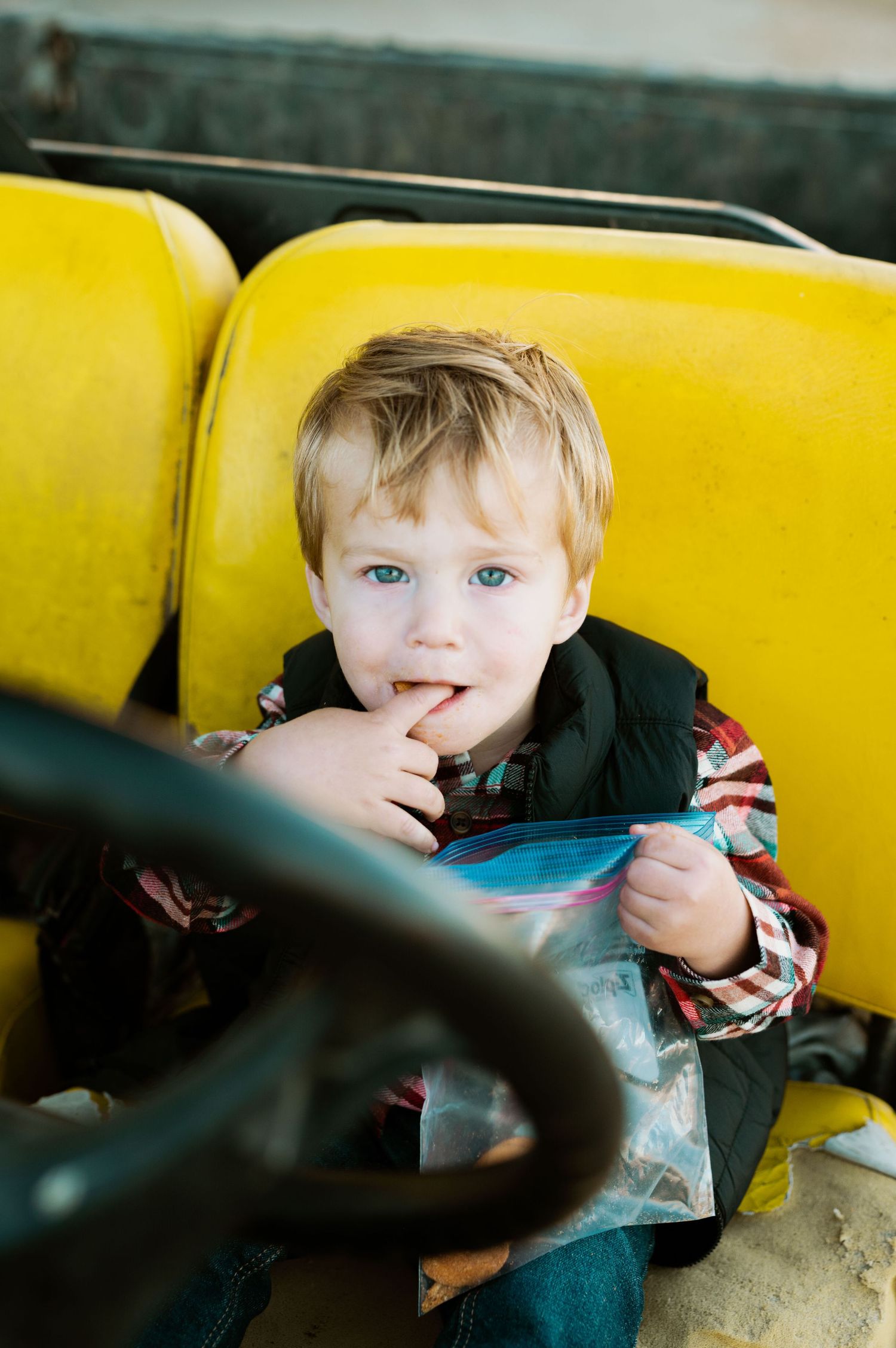 A small child sits in a yellow seat while holding a plastic bag and putting their finger in their mouth.