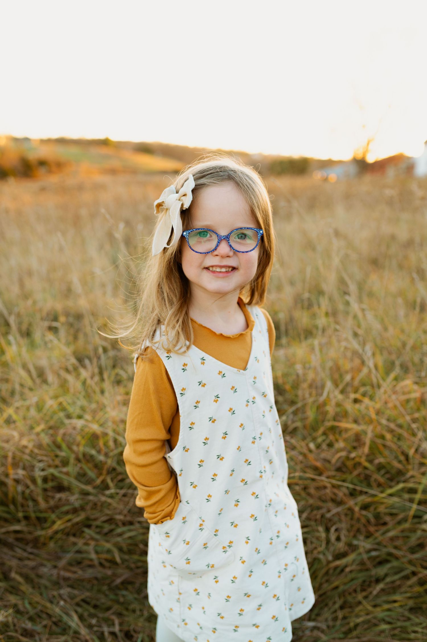 A child in blue glasses and a floral white dress stands in a golden field at sunset wearing a mustard yellow shirt.