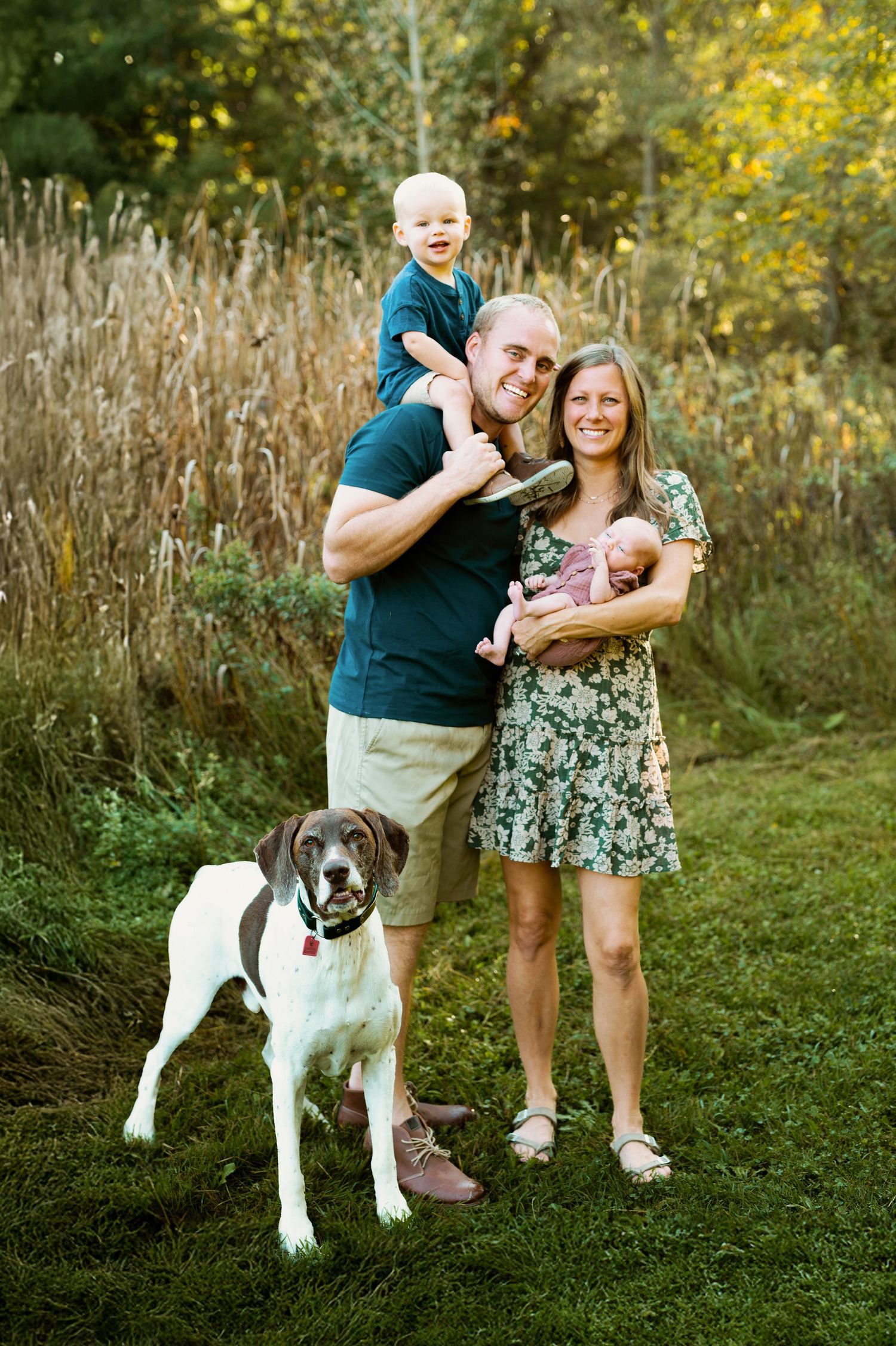 A couple and their black and white dog pose playfully in a grassy outdoor setting with golden sunlight.