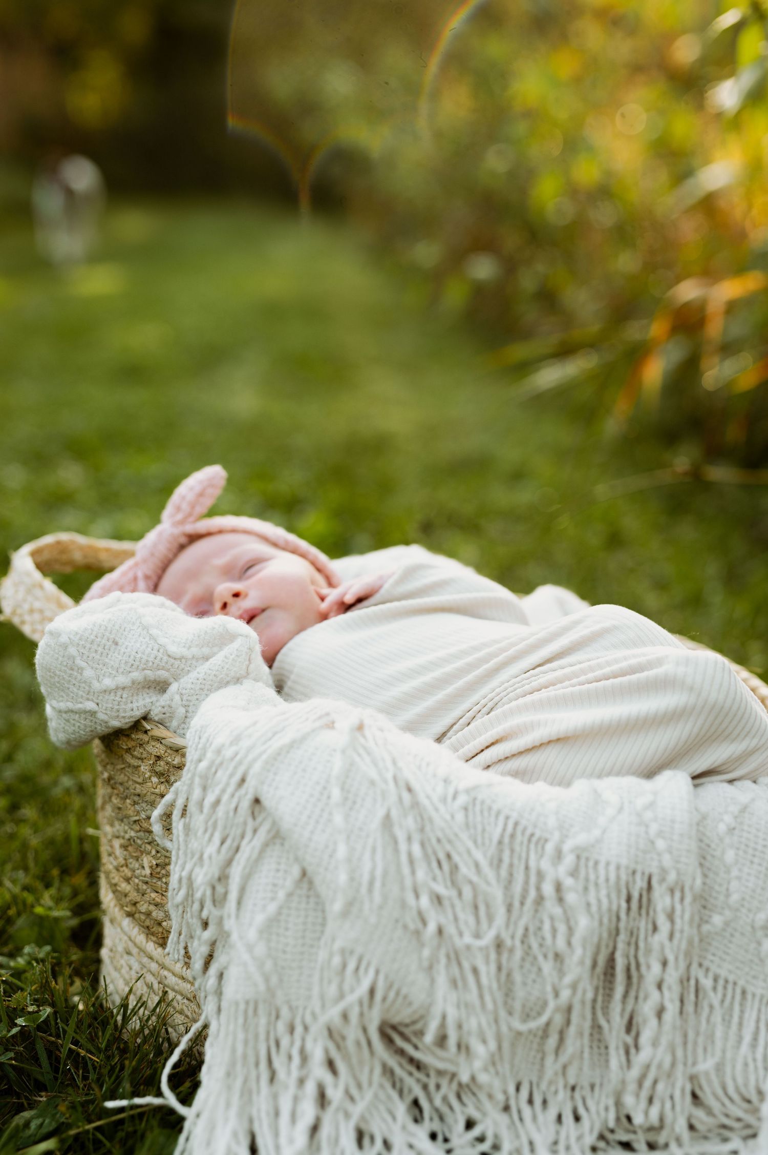 Newborn baby resting peacefully outdoors on white blanket with natural green background.