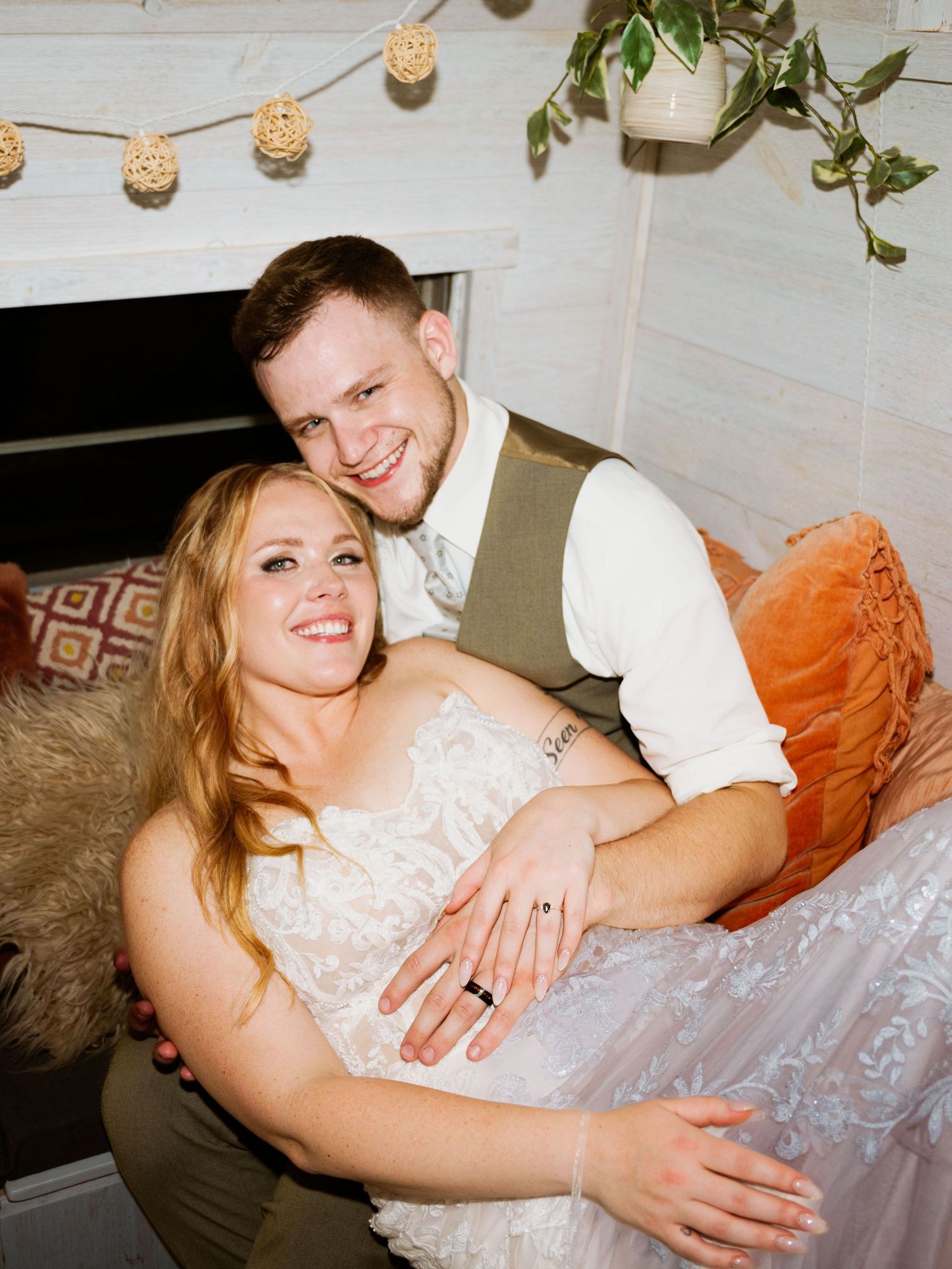 A relaxed wedding couple shares a candid moment on a cozy couch with string lights overhead.