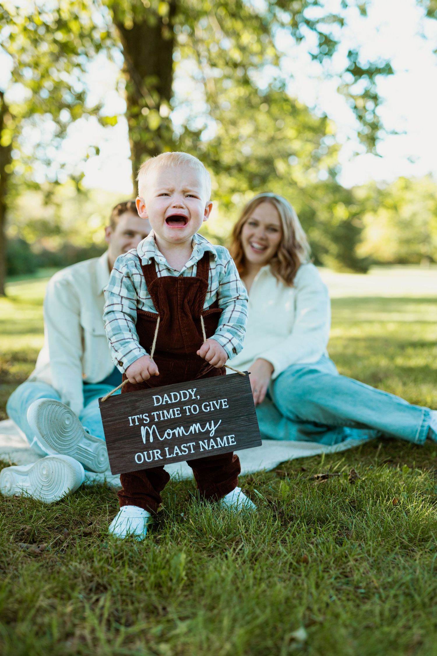 A laughing toddler holds a sign while sitting on grass with parents during a photo announcement.