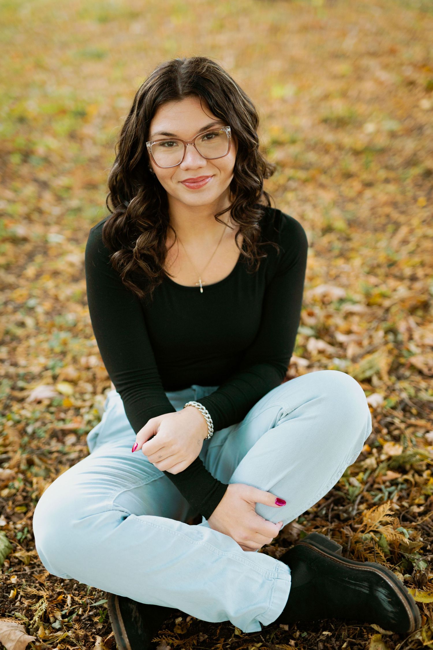 A person wearing glasses and a black sweater sits cross-legged on autumn leaves during an outdoor photoshoot.