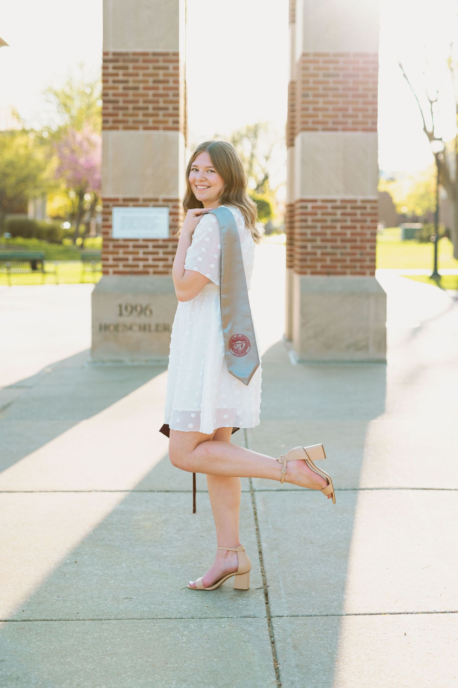 A graduate strikes playful pose by campus clock tower in white dress and stole.
