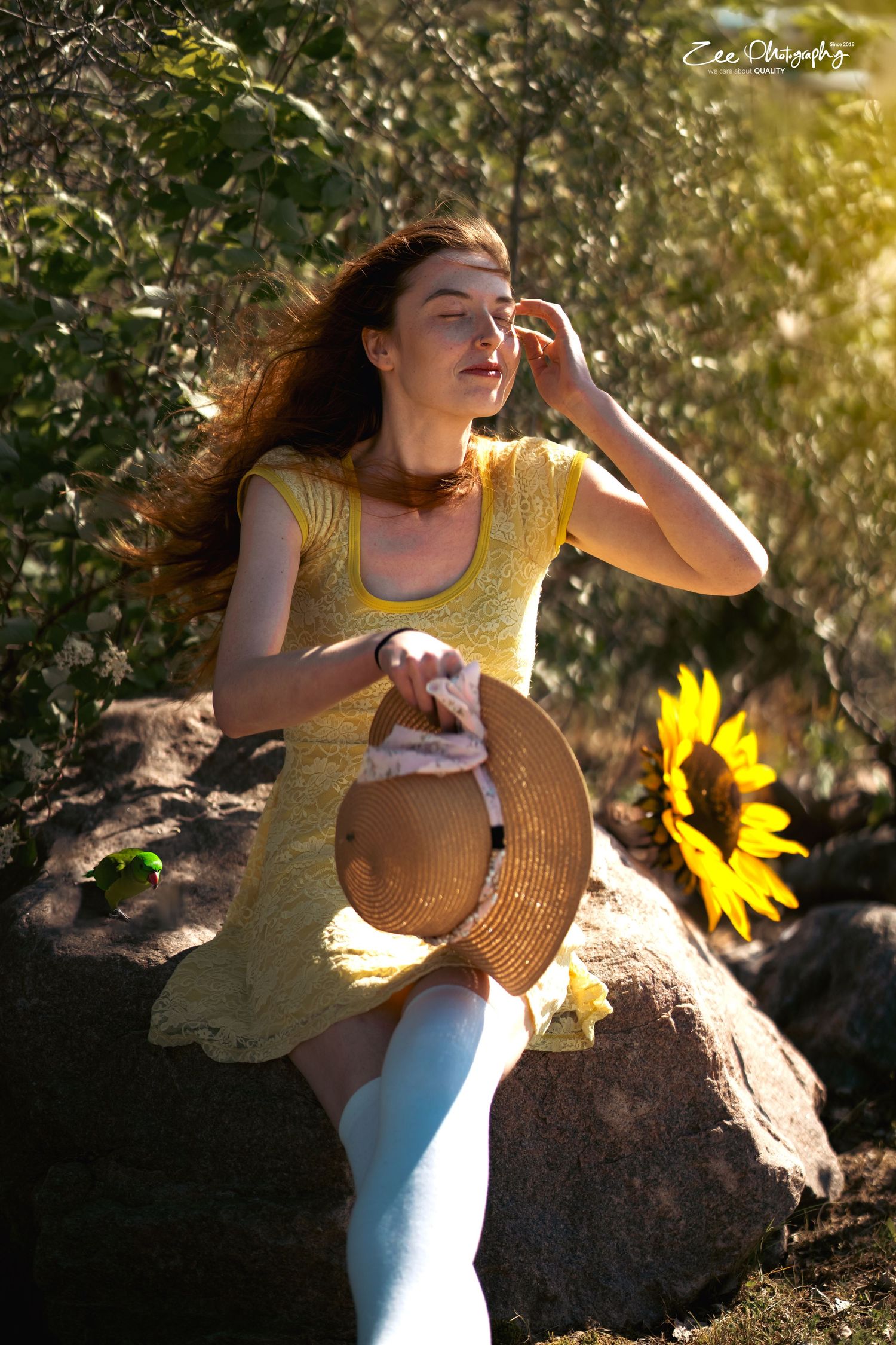 A person in a yellow sundress poses gracefully near a sunflower on a sunny afternoon, holding a straw hat.