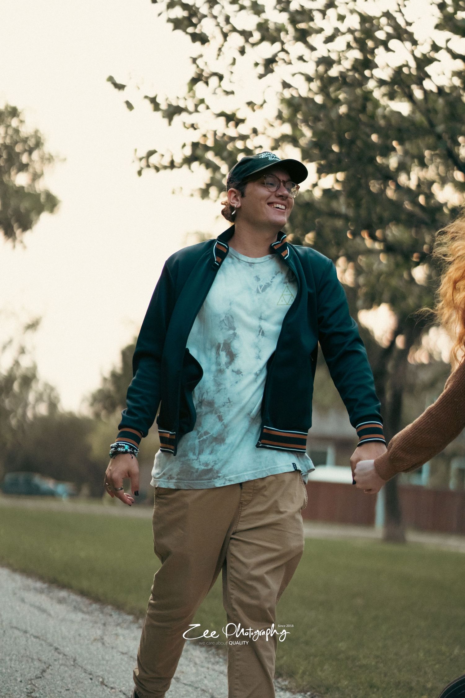 Person in navy jacket and baseball cap smiling while walking outdoors at sunset in park setting.