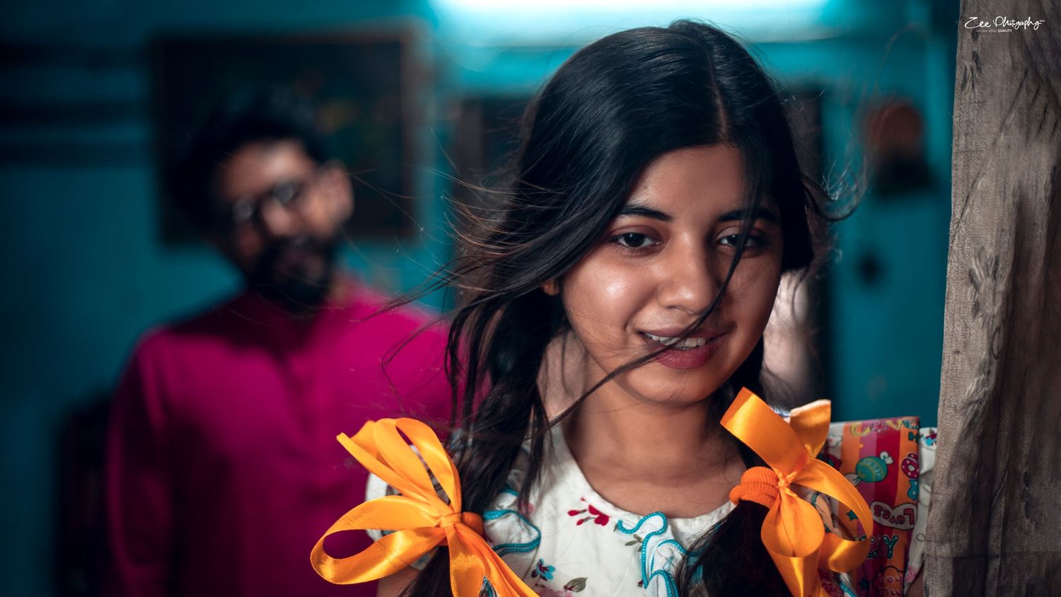 A person holding vibrant orange lilies smiles while posing against a teal background with someone in red visible behind them.