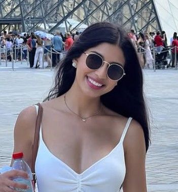 Tourist in white dress and sunglasses poses in front of glass pyramid structure at popular tourist destination.