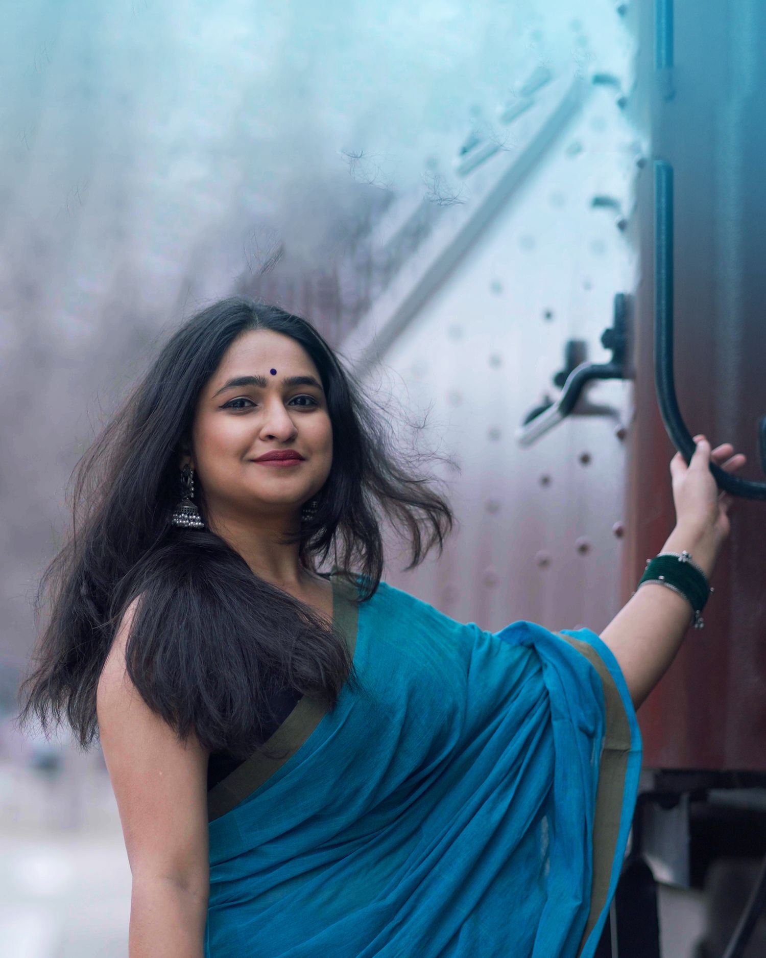 A person wearing a teal blue saree poses in front of a train car on a misty day.