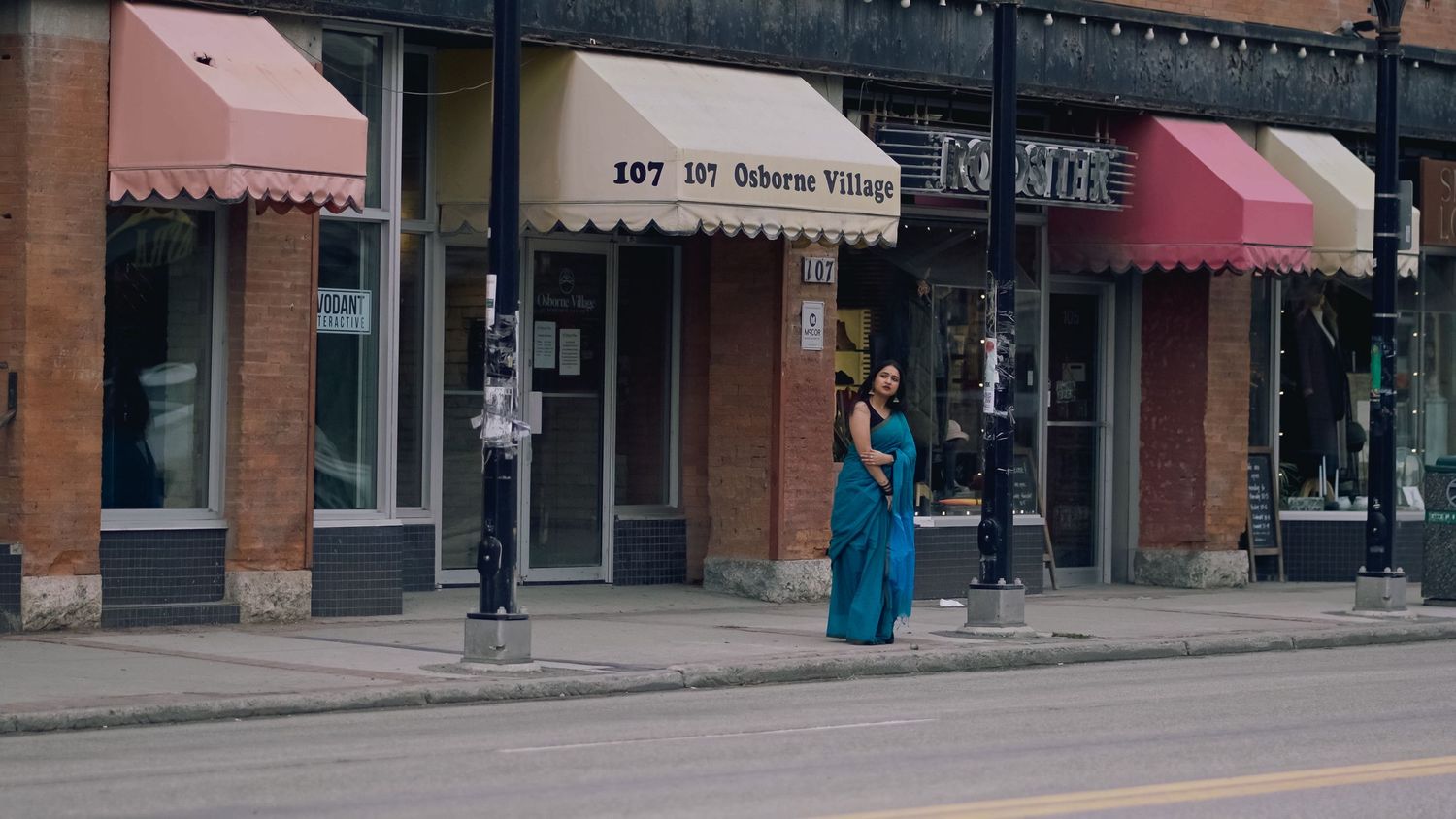 A person in a bright blue dress stands outside a row of storefronts with red awnings on a city street.