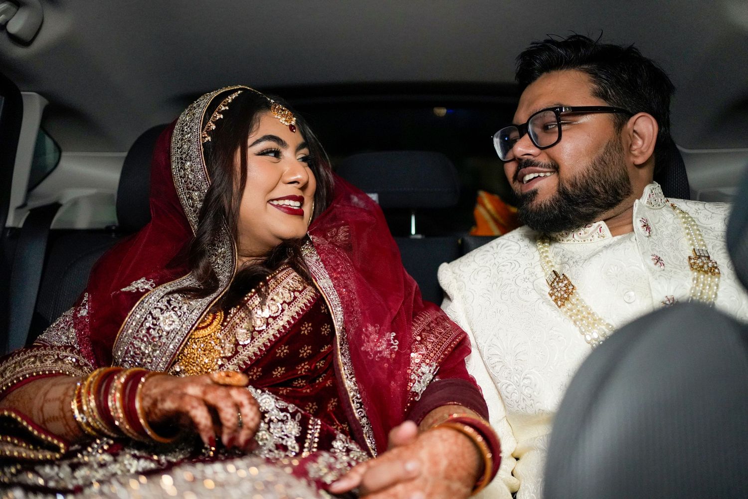 A couple in traditional South Asian wedding attire share a joyful moment while sitting in a car.