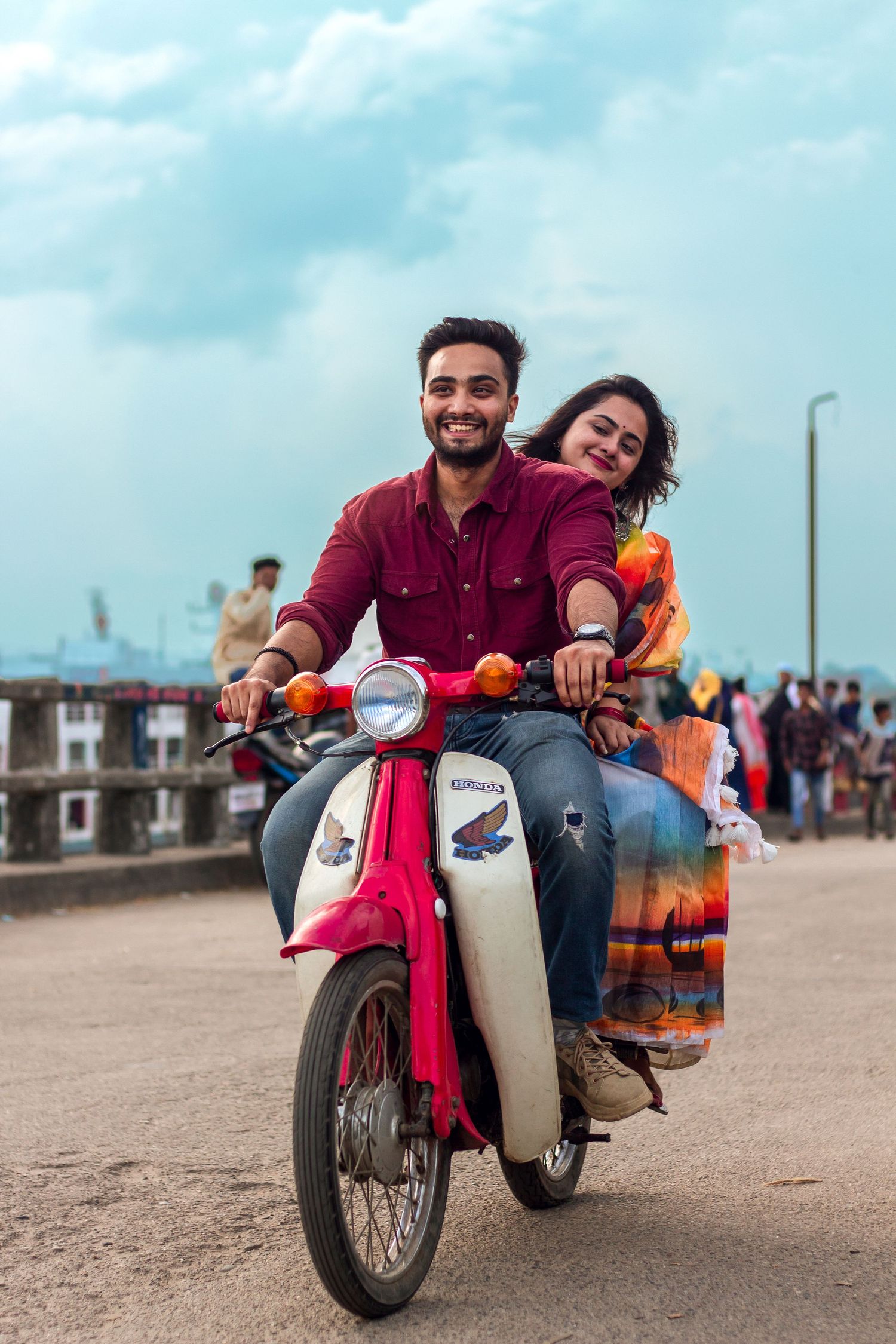 A couple riding a vintage red and white scooter along a seaside promenade on a sunny day.