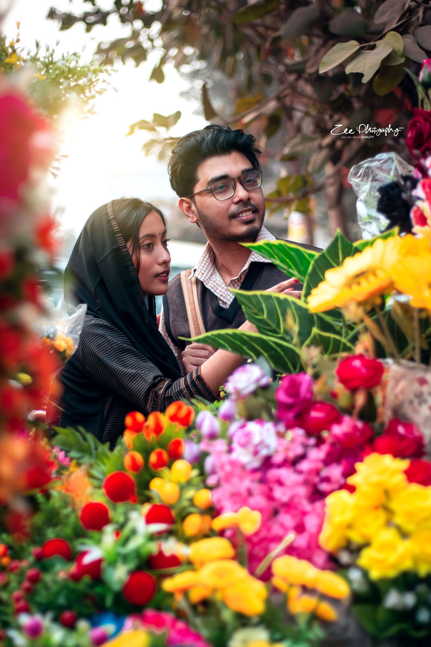 A couple admires colorful flowers including red, yellow and pink blooms in a garden setting with sunlit foliage.