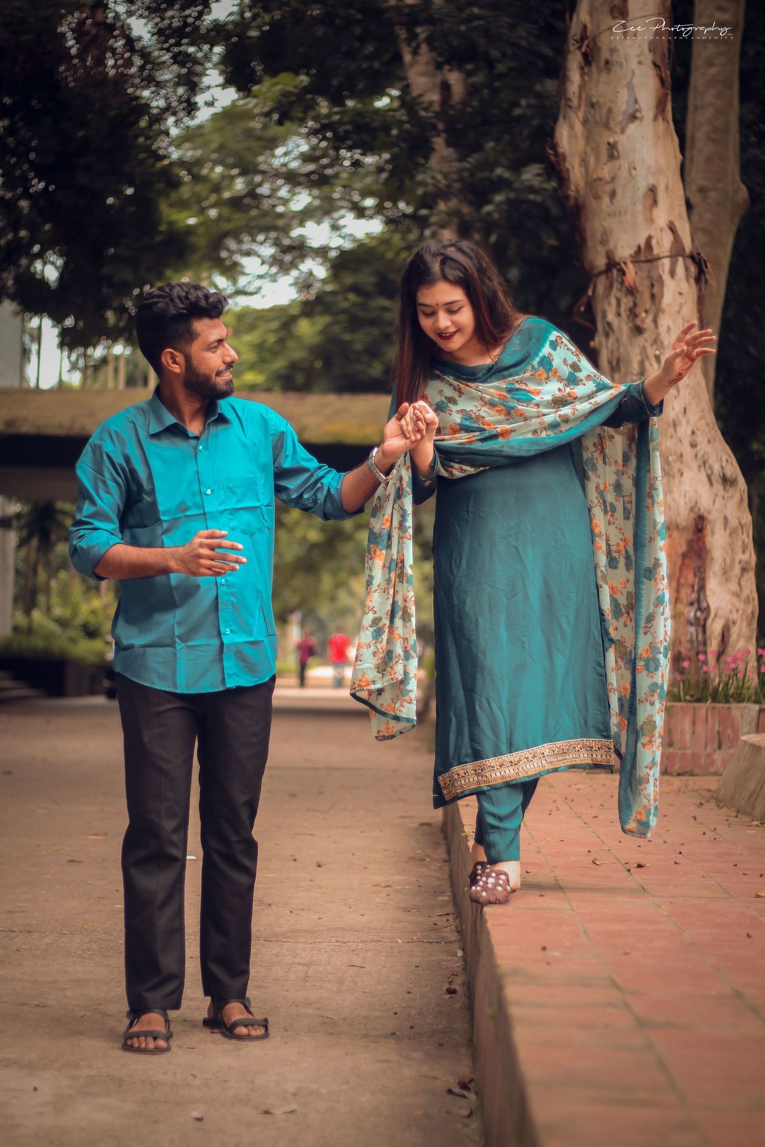 Couple in teal and floral attire creates candid moments on a brick pathway near trees.