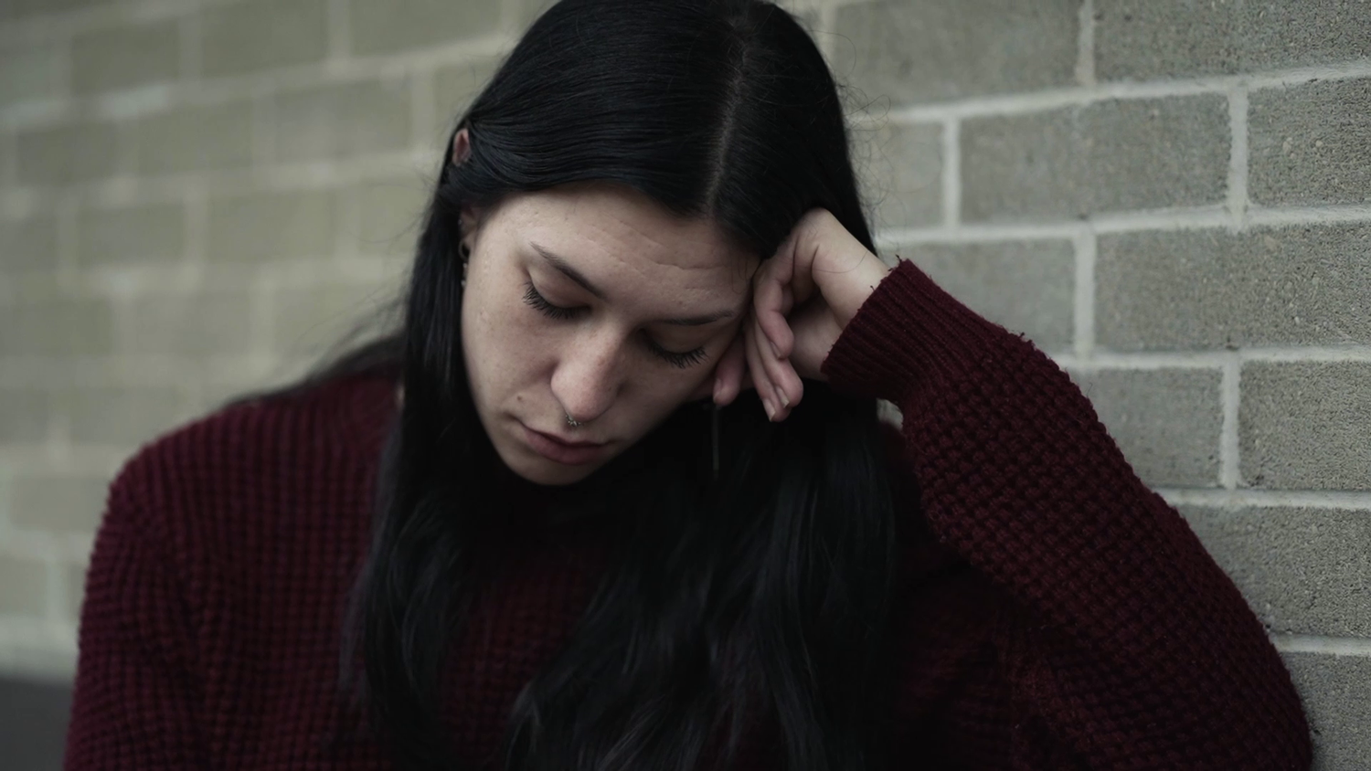 Woman in burgundy sweater sits against brick wall, touching forehead in distress.