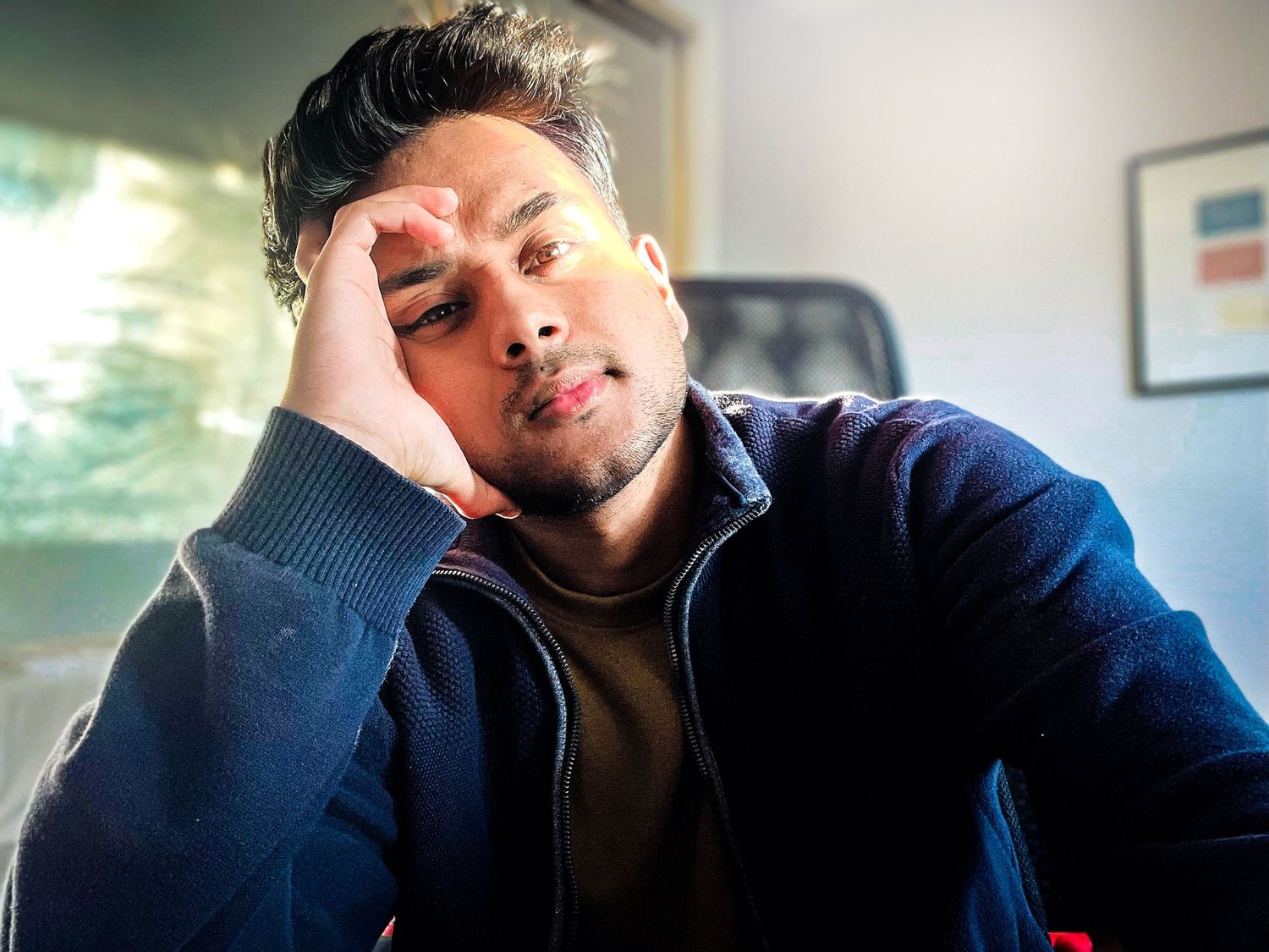 Person wearing a navy blue jacket and brown shirt rests their head on their hand while seated indoors.