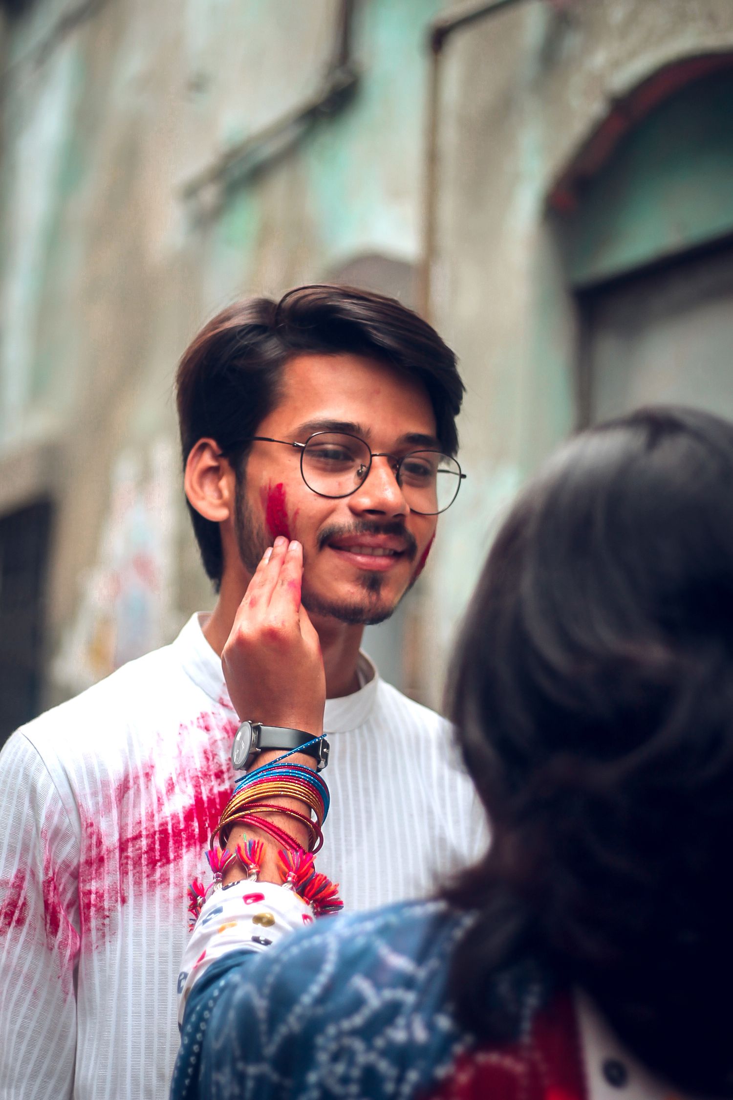 Someone with glasses and a white shirt gets colorful powder applied to their face during what appears to be Holi festival celebrations.