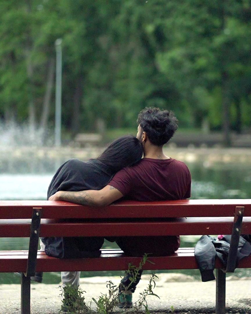 Two people sit close together on a red park bench overlooking a lake with lush greenery in the background.