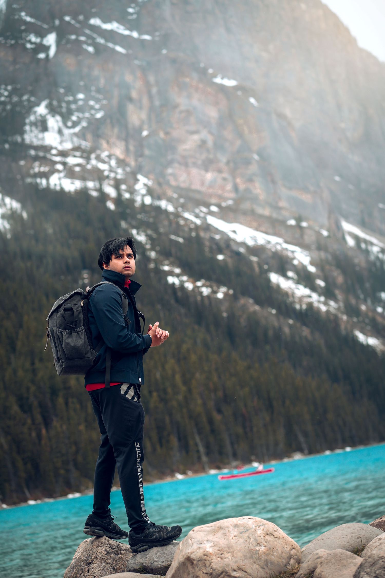 Hiker with backpack standing on rocks beside turquoise lake with snow-capped mountains.