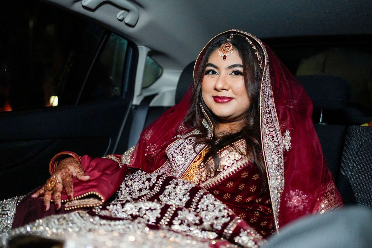 A traditional South Asian bride wearing an elegant red and silver bridal lehenga sits in a car.