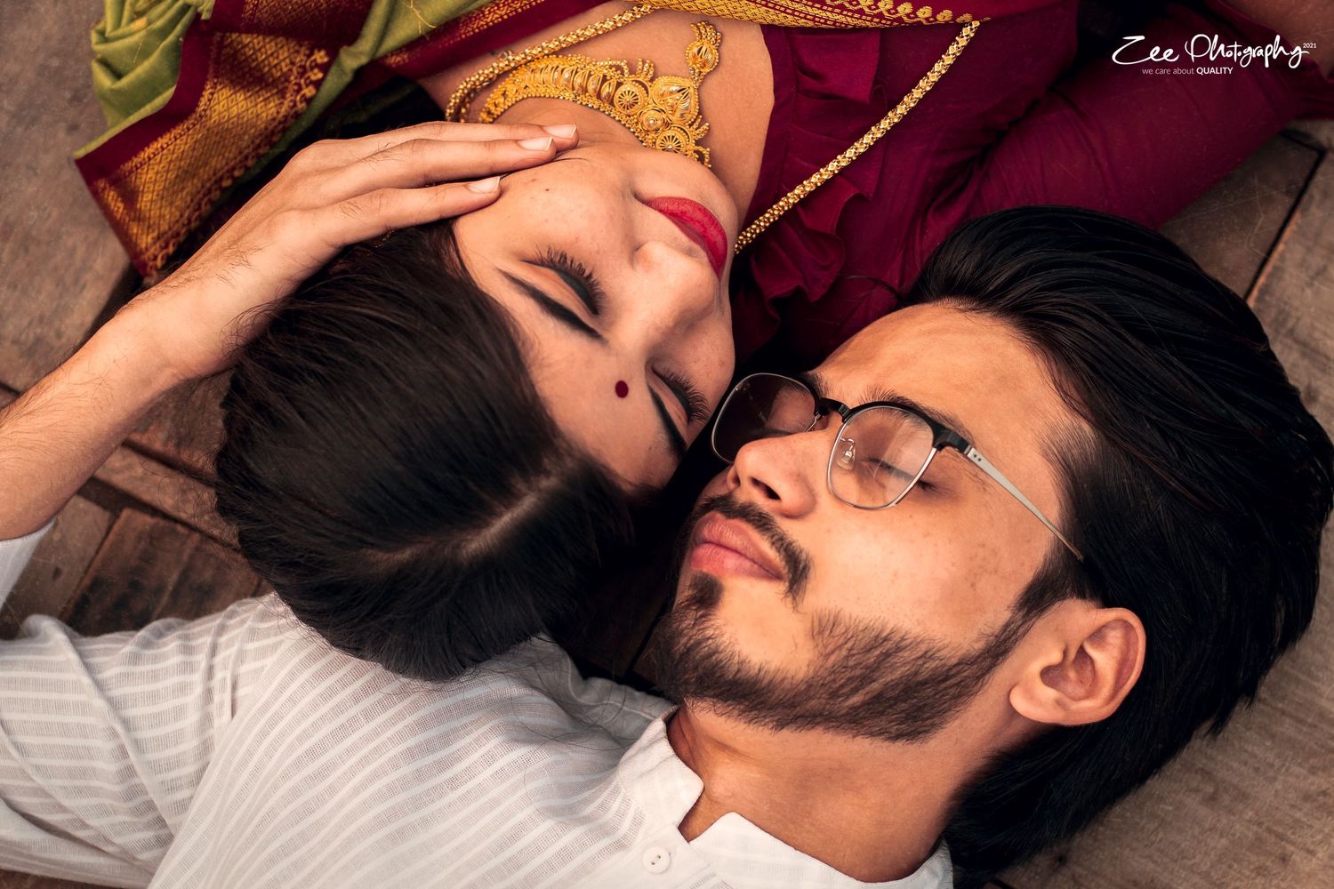 A couple lies together on wooden floor with one wearing a white shirt and glasses while the other rests peacefully.