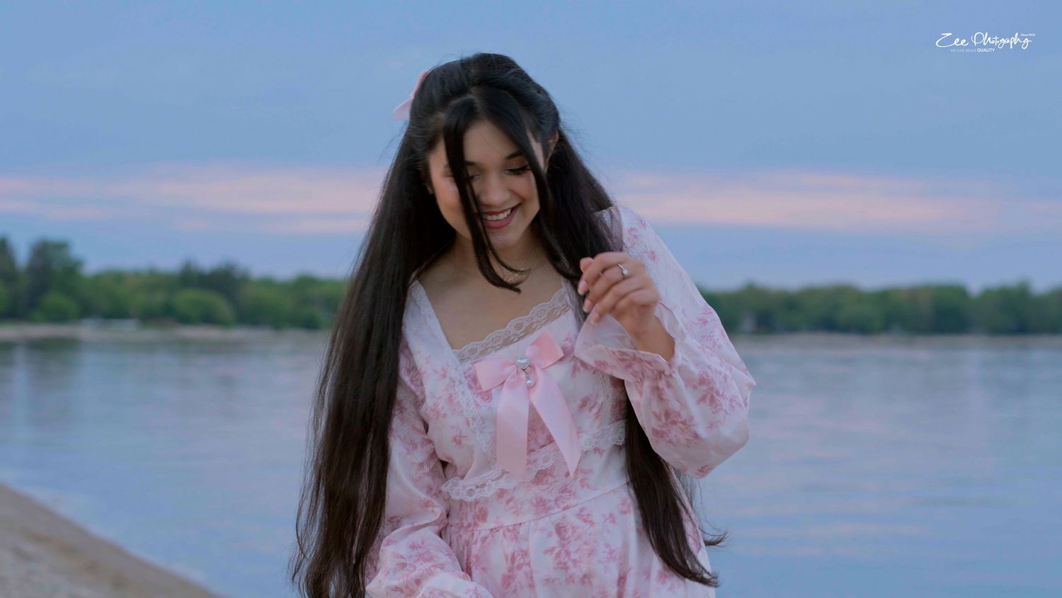 Long-haired woman in pink floral dress smiles while standing by tranquil lake at sunset.