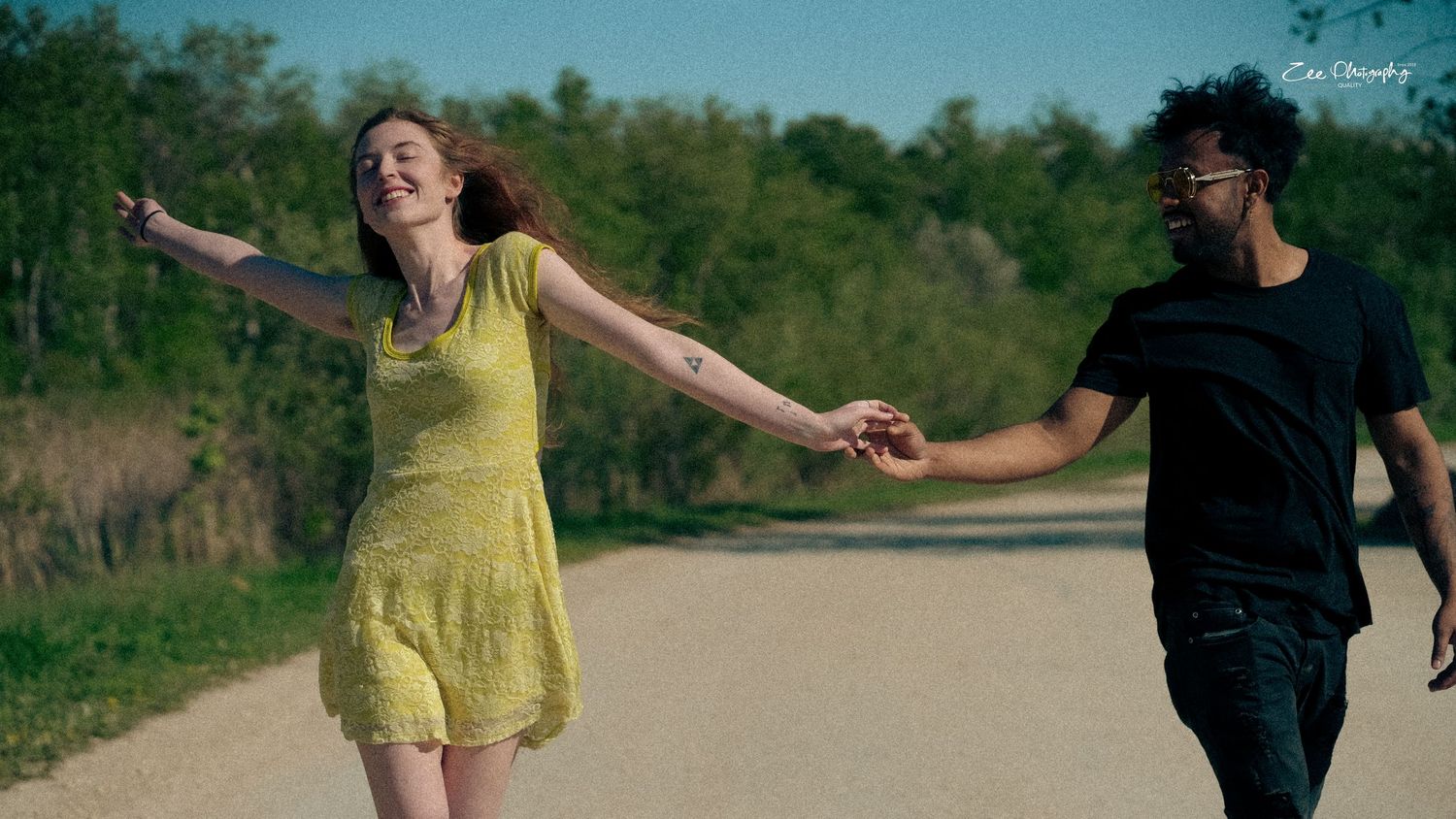 A couple playfully holds hands while walking on a rural road surrounded by green countryside.