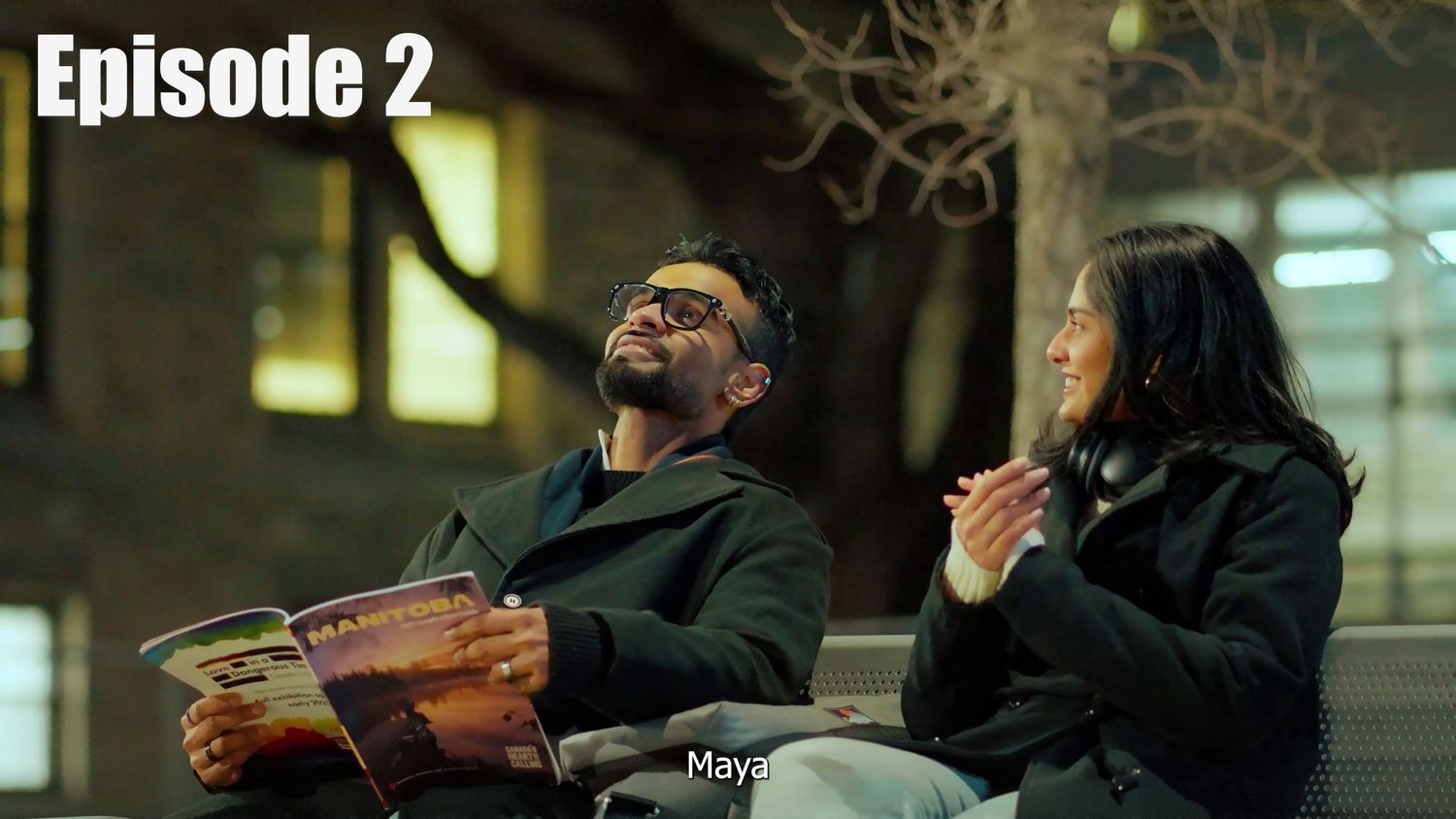 Two people sit together in a dramatic scene with low lighting, one holding an item while both appear emotional.