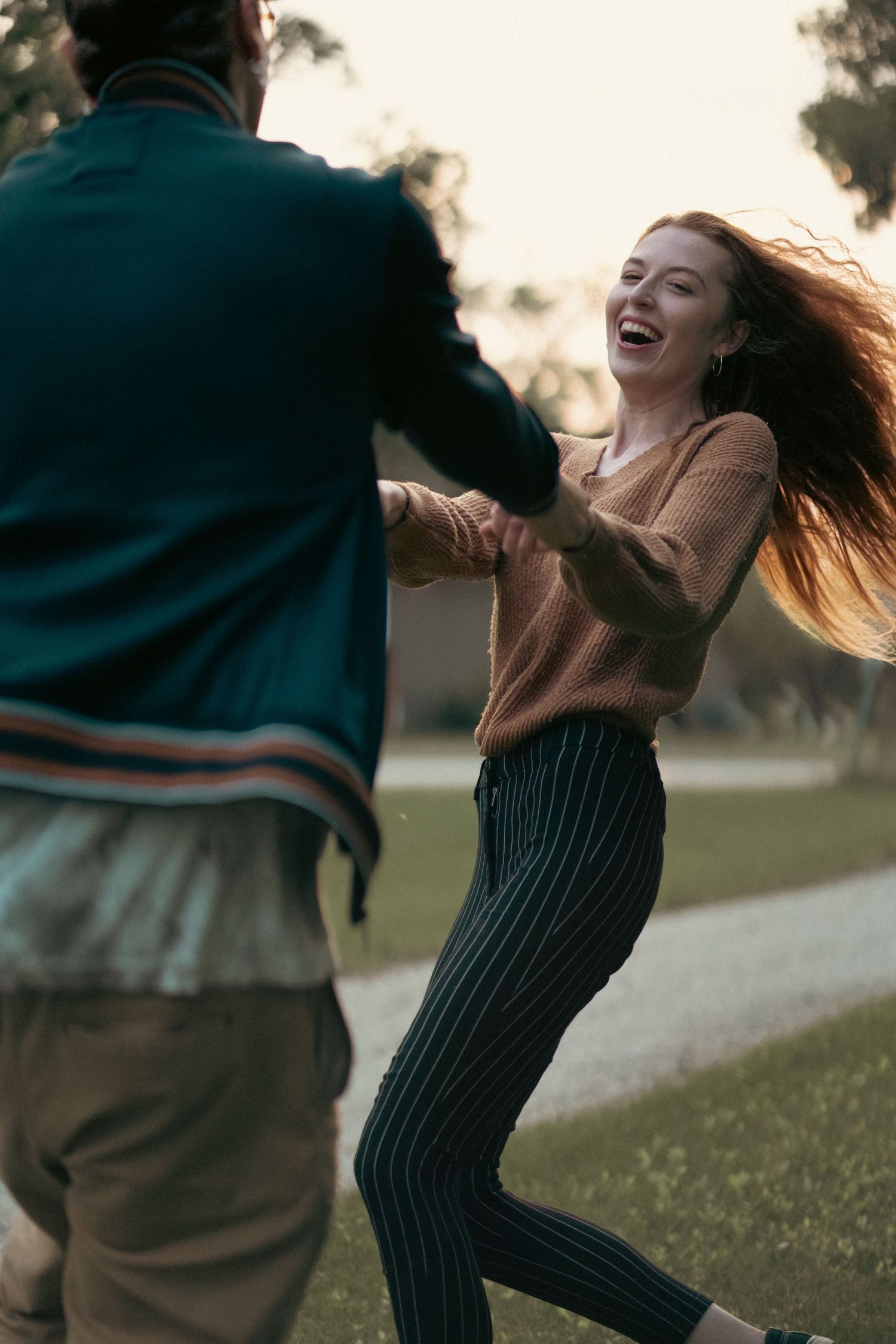 A couple dancing together in a park at sunset with one wearing a tan sweater and striped pants showing joyful movement.