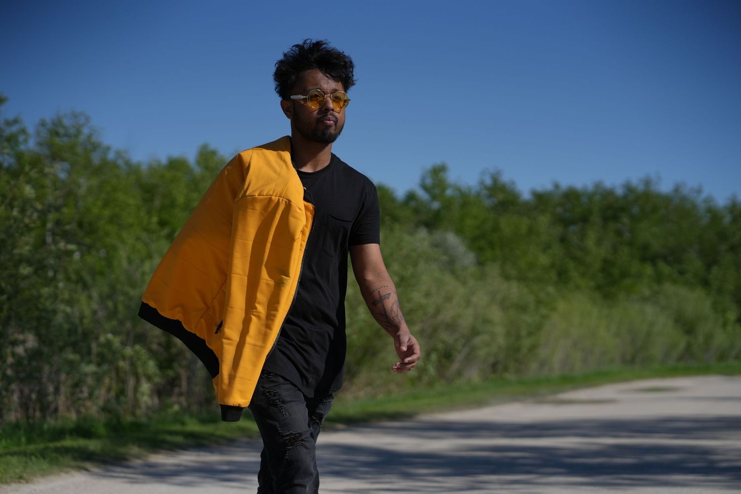 Person wearing a black shirt and holding a yellow jacket walks along a rural road on a sunny day.