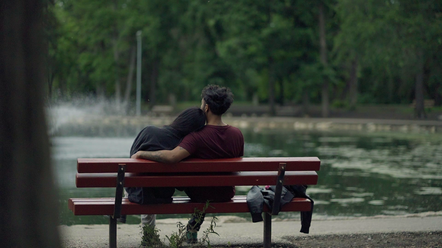 Someone sits alone on a red park bench overlooking a serene pond surrounded by green trees on a calm day.